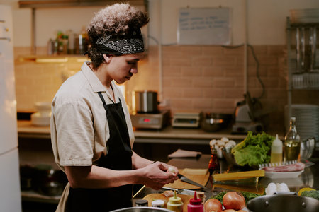 Young male cook peeling garlic to cook delicious meat while standing in modern professional kitchen of restaurantの写真素材