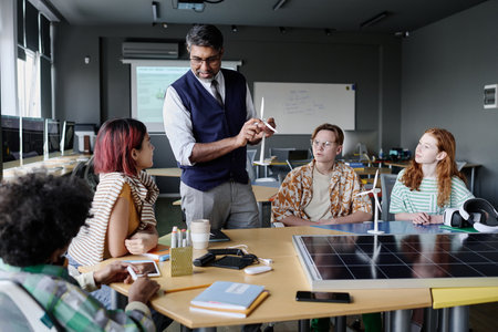 Mature biracial teacher working with group of modern teenagers telling them about wind power and...