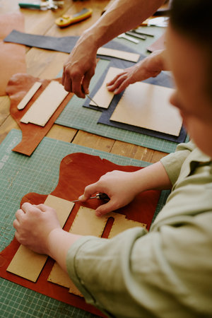 High angle shot of tailors hands cutting out and poking leather workpieces which lying on old wooden...