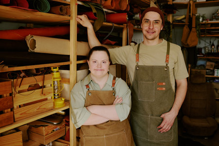 Male and female with down syndrome tailors standing next to wooden shelves full of leather rolls and...