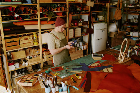 High angle shot of male tailor holding deep blue colored leather workpiece and looking at it at work...