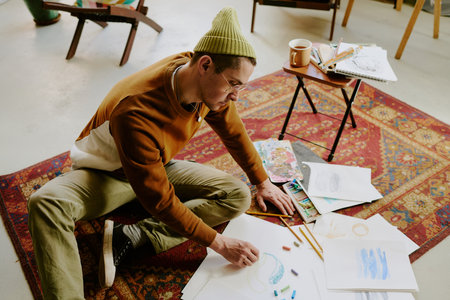 High angle view of young Caucasian man sitting on vintage carpet drawing picture on paper with crayonsの写真素材