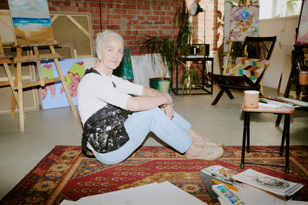 Flash shot of creative senior Caucasian woman sitting on vintage carpet on floor in her art studio and looking at cameraの写真素材