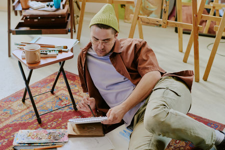 Young male artist wearing casual clothes drawing pencil sketch while relaxing on floor in his studioの写真素材