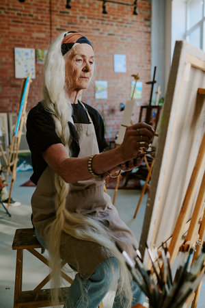 Vertical medium long shot of senior Caucasian woman with long gray hair sitting in front of easel and painting on canvasの写真素材