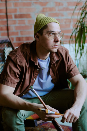 Vertical medium shot of young Caucasian male painter holding paintbrushes sitting in art studio and...