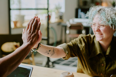 Young Caucasian female programmer with tattoo on arm doing high five with unrecognizable colleague...