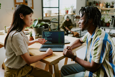 Young Black male and Asian female developers discussing code while their unrecognizable coworker...