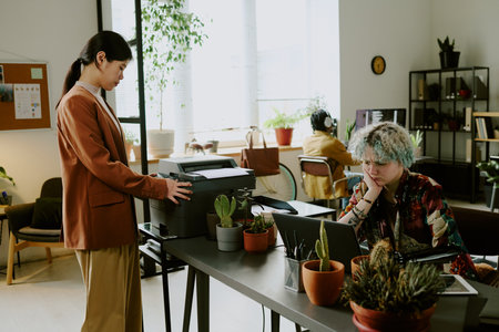 Young Asian woman printing documents while her Caucasian colleague searching for mistake in code at work in modern IT company officeの写真素材