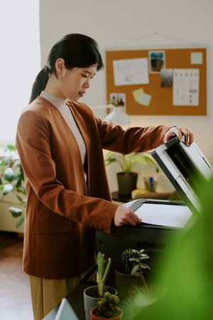 Vertical medium long shot of Asian female manager making copy of paper document with use of scanner in modern officeの写真素材