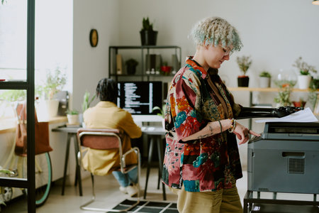 Young gen Z woman with prosthetic arm using printer in modern office, her male colleague typing code...