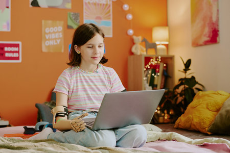 Teen girl with disability wearing prosthetic arm sitting on bed in her bedroom and using Internet...