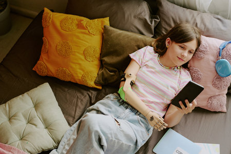 From above view of modern Caucasian teen girl with disability relaxing on comfortable bed and surfing Internet on smartphoneの写真素材