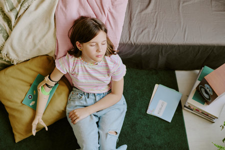 From above view shot of tired teen girl with disability sitting on floor in her bedroom and leaning...