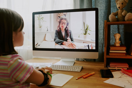 Modern teen girl with disability sitting at desk in her room listening to teacher during online...