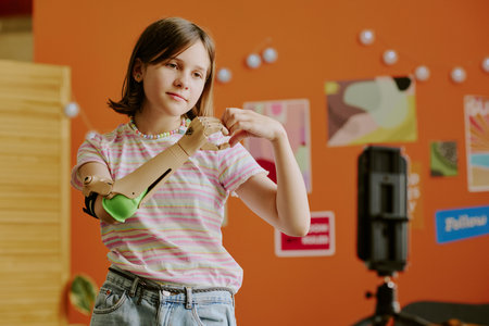 Teen girl with arm prosthesis standing in front of smartphone on tripod showing hand heart gestureの写真素材