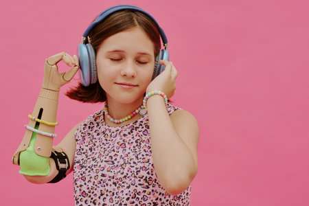 Medium studio portrait of happy teen girl with prosthetic arm listening to music in headphones, pink backgroundの写真素材