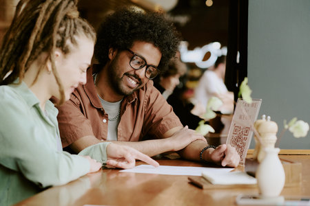 Smiling man with glasses and woman with dreadlocks discussing menu options in cozy cafe setting,...