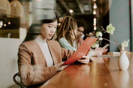 Asian woman reading menu at contemporary cafe interior with group of diverse individuals in background, sitting at wooden bar counter with plants in ceramic vasesの写真素材