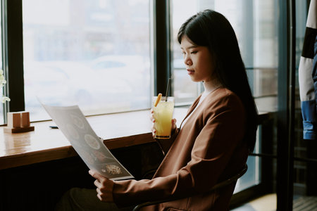Woman in brown blazer sitting by window, holding glass of lemonade and reading menu, in intimate...
