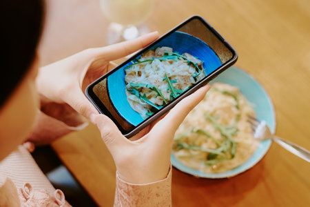 Person taking photo of delicious creamy pasta dish on table with smartphone, neatly garnished with...