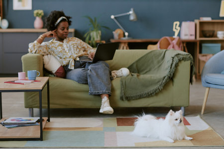 Woman sitting comfortably on couch using laptop with pensive expression in cozy living room setting and a small dog relaxing on floorの写真素材