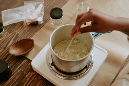 Person stirring wax in a metal pot on electric burner surrounded by jars and wooden spoon on wooden...