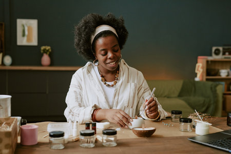 African American woman smiling while mixing skincare ingredients at wooden table cluttered with jars and tools in cozy home settingの写真素材