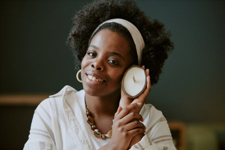 Woman with afro hairstyle and white headband smiling while holding a candle against her face,...