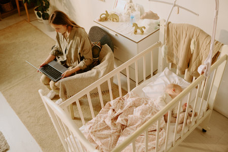 Young woman sitting on chair and working on laptop near baby crib with sleeping baby in cozy room filled with soft light creating relaxing atmosphereの写真素材