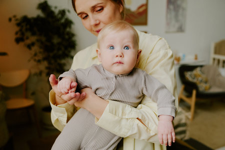 Portrait of mother holding her baby while standing in living room. Baby looking at camera with blue eyes and wearing striped outfit with soft lighting from windowの写真素材