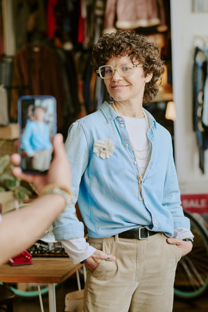 Woman with curly hair and glasses posing in vintage store. Smiling warmly while engaging with another person holding smartphone, creating friendly atmosphere in cozy shop environmentの写真素材