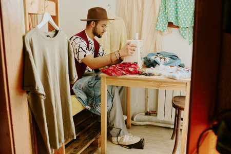 Young adult sitting in small workspace, surrounded by clothing and fabrics, working on new fashion design using creativity and artistic skillsの写真素材