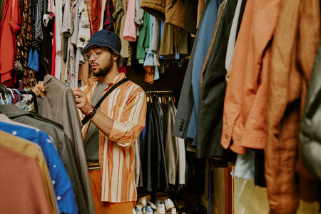 Young man with glasses and hat selecting clothes in a vibrant store with many options on both sides...