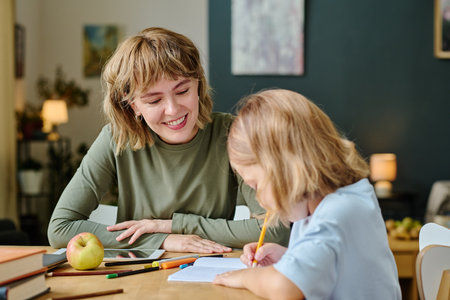 Young mother with bob haircut smiling widely and admiring her little girls art while she drawing