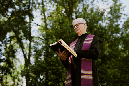 Low angle shot of senior priest in black vestment holding Bible and reading prayerの写真素材