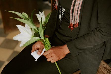 Unrecognizable woman dressed in black clothes sitting on wooden bench and holding white napkin with white lily during funeralの写真素材