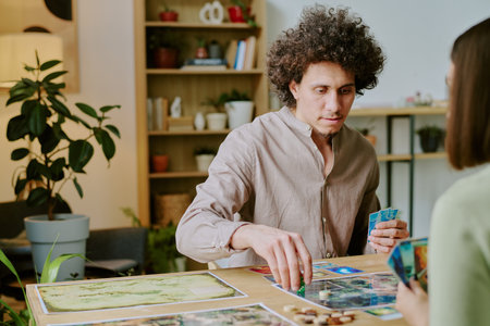 Young biracial man with curly hair sitting at table in living room and playing board game with unrecognizable girl friendの写真素材