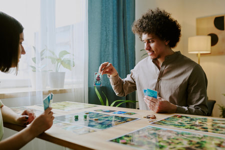 Young biracial man showing his card to girlfriend while playing modern fantasy board game together at homeの写真素材