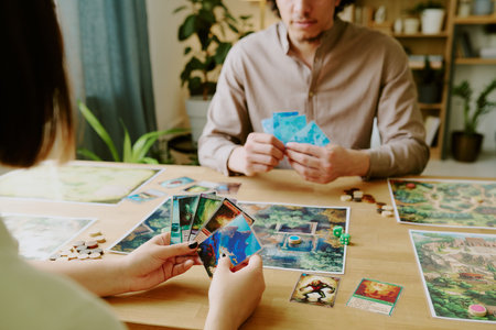 Unrecognizable young woman and her friend sitting at table in front of each other with cards in hands playing modern board gameの写真素材