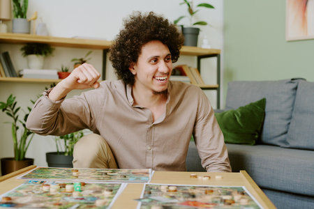 Medium shot of joyful young biracial guy sitting at coffee table in living room winning modern board gameの写真素材