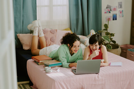 Two young women studying and discussing lesson material with laptop in cozy bedroom setting, lying on bed. Surrounded by books, plants, and photos on wallの写真素材