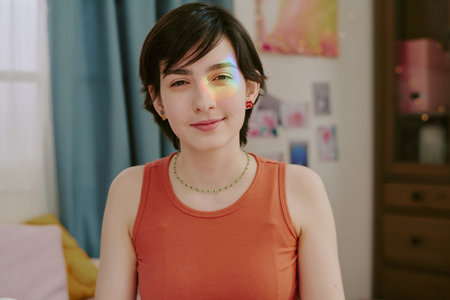 Portrait of young woman with short hair wearing an orange top and green necklace, sitting in a room with colorful decorations and blue curtains behind herの写真素材