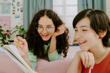 Two young girls are sitting close, sharing a story and smiling warmly. Room is decorated with plants, photos, and colorful items, creating a cheerful ambianceの写真素材