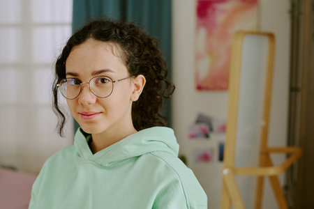 Portrait of young woman wearing glasses and casual hoodie, smiling softly while looking straight at camera. Scene includes a background with blurred mirror and artworkの写真素材