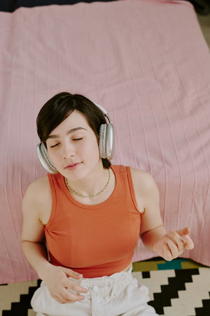 Woman wearing headphones enjoying music while sitting on a bed with a calm expression on her face, bedspread and patterned rug visible in backgroundの写真素材