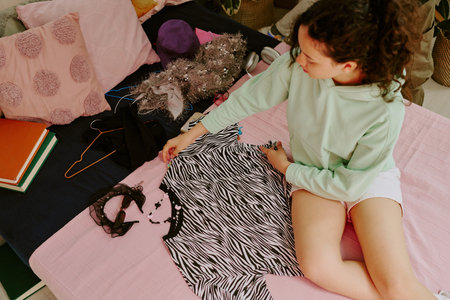 Young girl with long curly hair selecting zebra-print shirt on bed filled with other clothes and...
