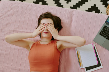 Person lying on pink blanket with hands covering face, adjacent to laptop and notebooks on patterned rug, creating a scene of relaxation and possible study sessionの写真素材