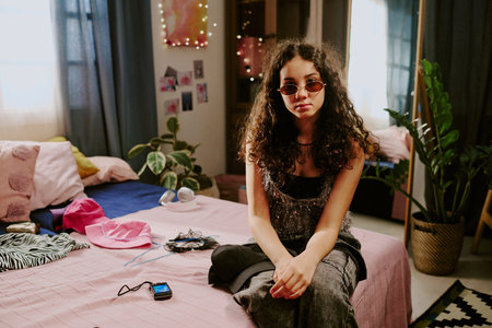 Portrait of a young woman with curly hair wearing sunglasses, sitting on bed in cozy room with personal decor and plants around, looking confident and relaxed at cameraの写真素材