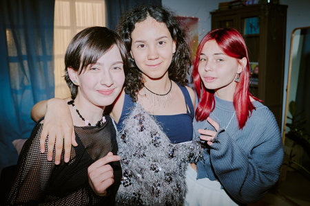 Three young friends standing close together, smiling warmly, with two looking directly at camera. Background includes indoor setting with curtains and furnitureの写真素材